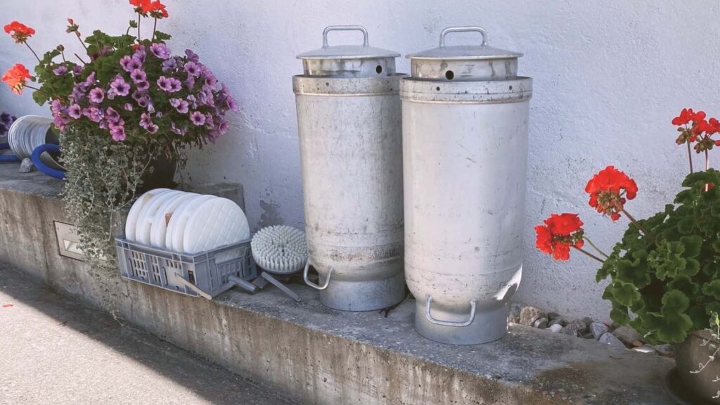 Zwei alte Milchkannen vor einer Käserei, umrahmt von Blumen – Sinnbild für Wurzeln, Übergänge und behutsame Veränderung