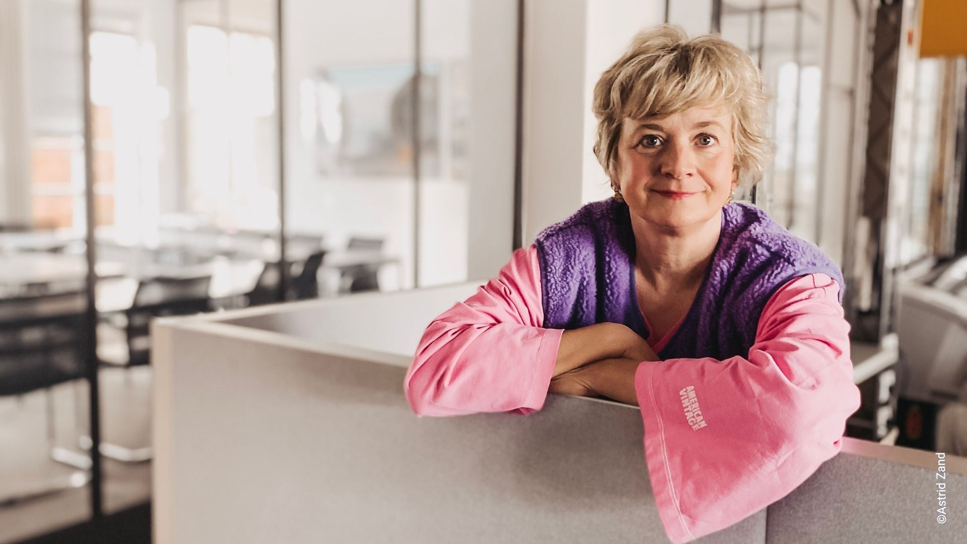 Astrid Zand lehnt lächelnd an einer Trennwand in einem Großraumbüro und schaut in die Kamera
